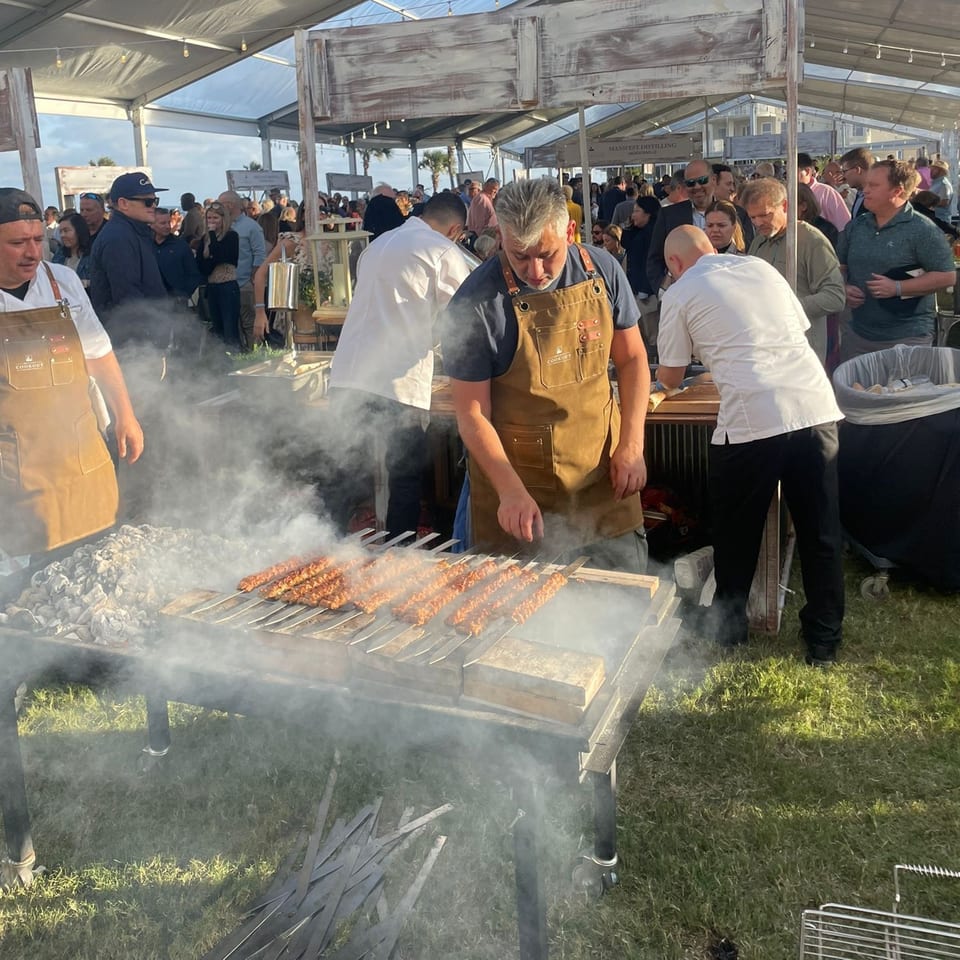 Chefs grill Turkish kebabs on a smoky outdoor grill under a tent, surrounded by people.