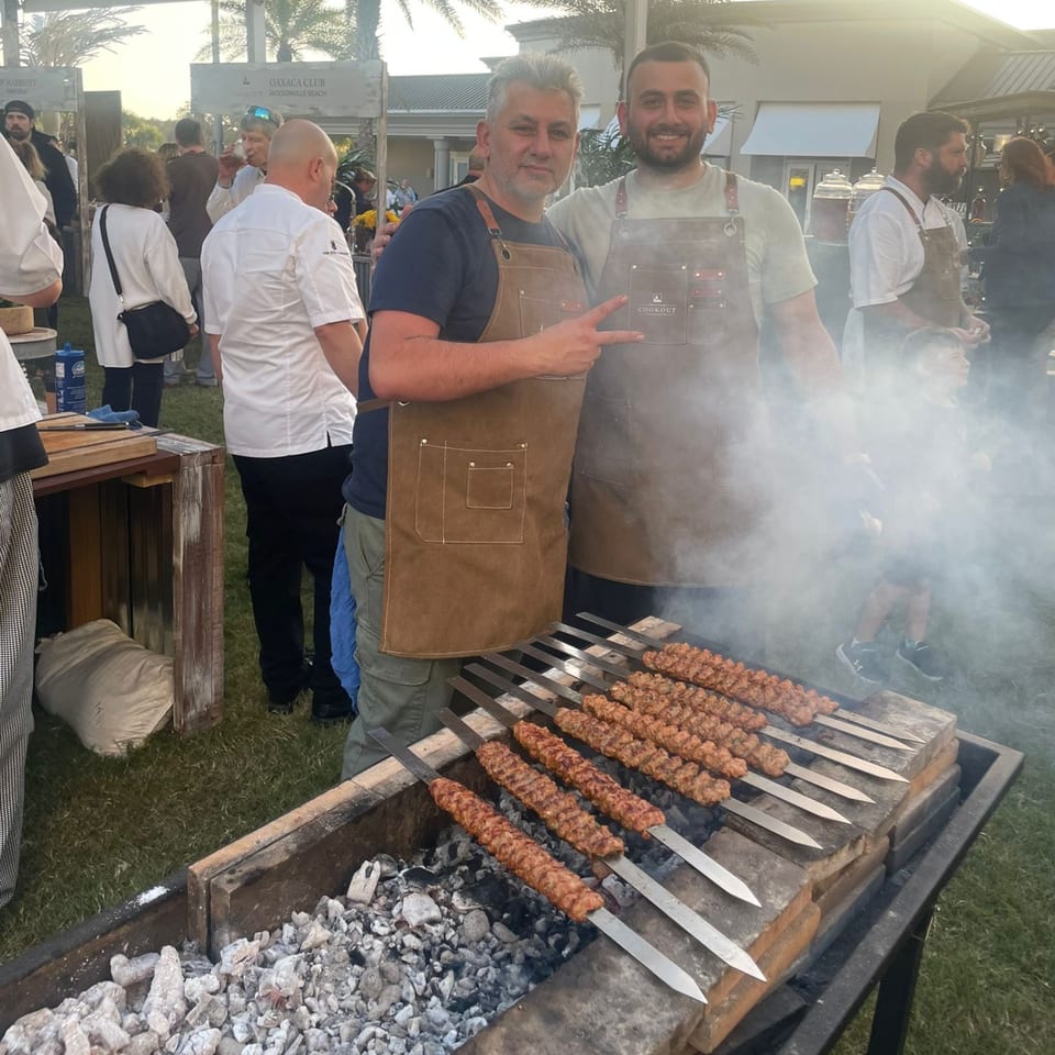 Two men in aprons pose by a grill with Turkish Adana kebabs cooking over coals, smoke rising.